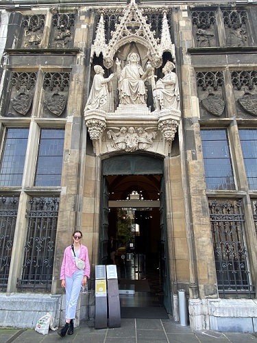 Orla stands outside the wooden door of a church, smiling and wearing sunglasses. Stone sculptures of saints are above the large wooden door.