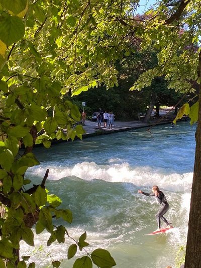 A surfer on the man-made surf in the Englischer Garten in Munich. It is a sunny day, and spectators watch from the banks.