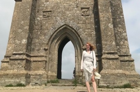 Alice stands at the base of Glastonbury Tor. She is smiling and the wind has caught her hair. Behind her, it is possible to see from one side of the Tor to the other through the stone archways.
