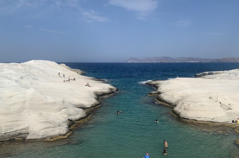 Two outcrops of Sarakiniko beach, which is formed from white volcanic rock rather than sand. In channel between the two outcrops, there are people swimming in the shallow, turquoise waters. In the background, the sea becomes a deeper blue and the outline of a distant island can be seen.