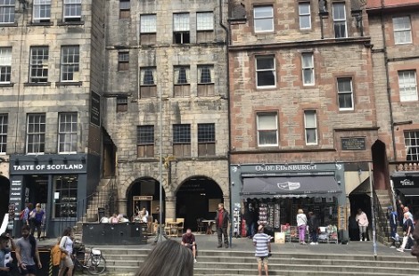 The photo shows the Royal Mile in Edinburgh, with tall stone buildings lining the street. Some people are handing out flyers for performances. Two shops on the street are called 'Olde Edinburgh' and 'Taste of Scotland'.