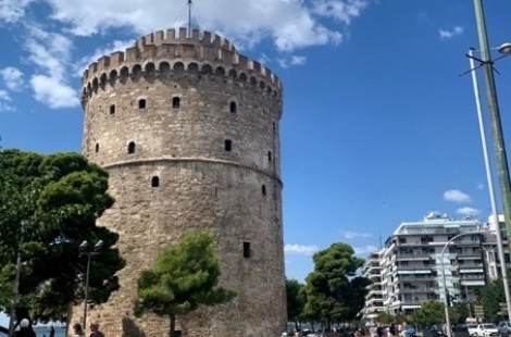 A photograph of the White Tower on Thessaloniki, which is built in light stone and dominates the landscape. It is a sunny day, with people gathered around the base of the tower.