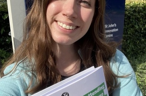Helen stands smiling in front of a St John's Ambulance sign, holding a pack of documents for her course on Mental Health First Aid.