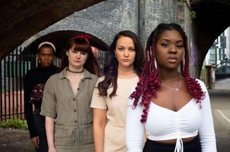 Joy stands with three other poetry performers looking directly into camera. The group is stood under railway arches in Manchester. Joy is wearing a white top and has red braids.