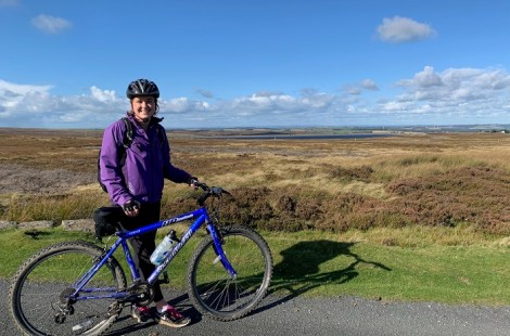 Lauren stands smiling with her bike overlooking the moor near Consett. She is wearing a purple waterproof coat and bike helmet. There is bright sunshine and few clouds in the sky.