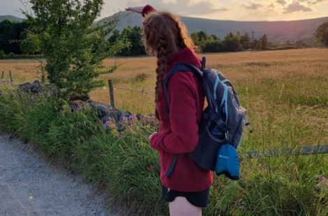 Nicole stands facing the peak behind her, playfully measuring the mountain with her hand. She is wearing a read fleece and is carrying walking gear. The sun is setting, so the light is very golden across the field of wheat in the valley.