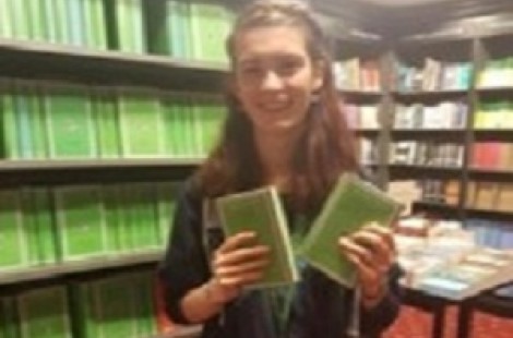 Ruth stands smiling in a book shop, holding two Latin books. The books have distinctive green covers on them, and there are many shelves of these green books behind where she stands.