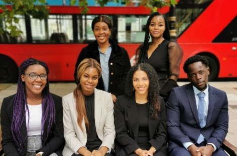 Sikemi smiles with a group of five other interns on the BUILD @ Sherman & Sterling Scheme. They are in business attire and are grouped in front of a red London bus.
