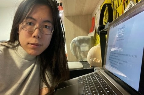 Xin looks into the camera smiling and wearing a pair of glasses. She is next to her laptop for the summer school.